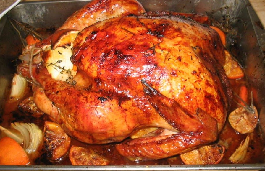 A whole wet-brined turkey roasted to a glossy, deep mahogany brown in a metal roasting pan, surrounded by roasted orange and lemon halves, onion wedges and thyme sprigs