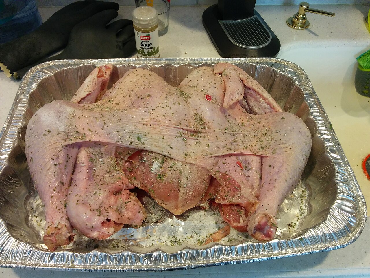 A raw butterflied turkey lying skin-side up in a disposable foil roasting pan, sprinkled with coarse salt and dried green herbs, with a jar of ground sage behind it