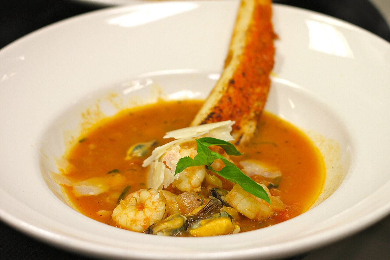 White bowl of tomato seafood stew with shrimp and mussels, topped with basil and a long toasted crouton