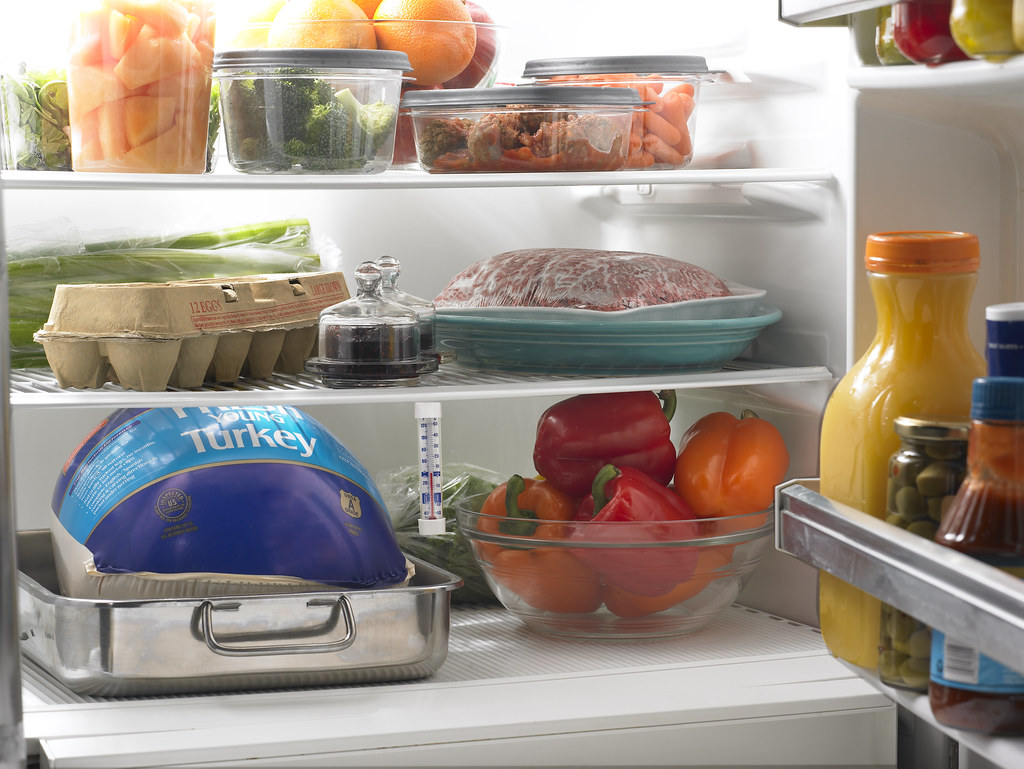 A frozen whole turkey still in its wrapper, sitting in a metal pan on the bottom shelf of a refrigerator with a fridge thermometer beside it