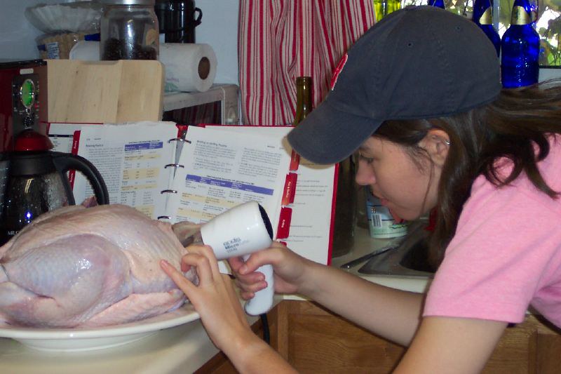 A person in a baseball cap aiming a hair dryer at a raw turkey on a plate, with a cookbook open behind it