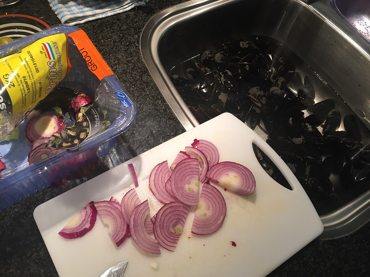 Raw mussels soaking in a sink of cold water next to a cutting board of sliced red onion