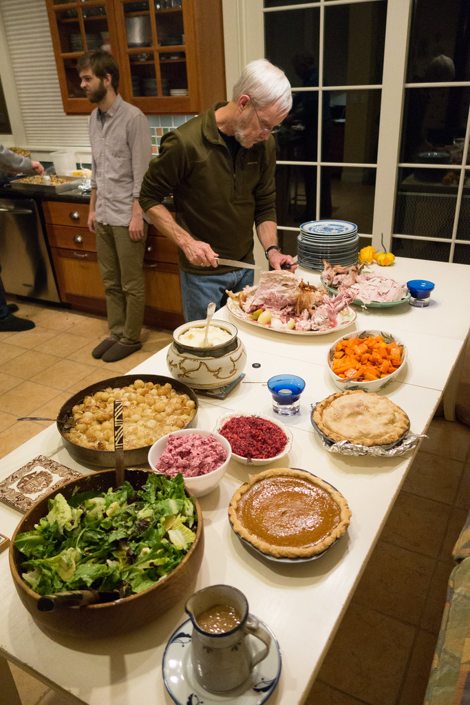 A man carving a turkey at the end of a kitchen buffet with stuffing, sweet potatoes, cranberry sauce, salad, gravy and two pies