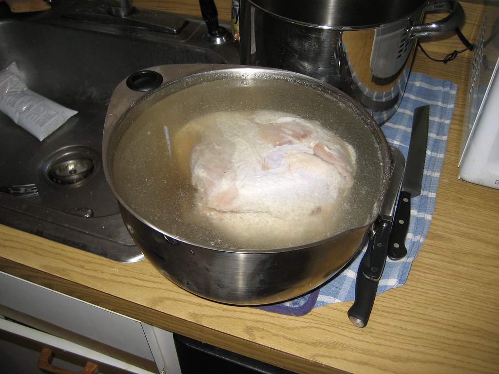 A whole raw turkey sitting in a large stainless steel bowl filled with water inside a kitchen sink