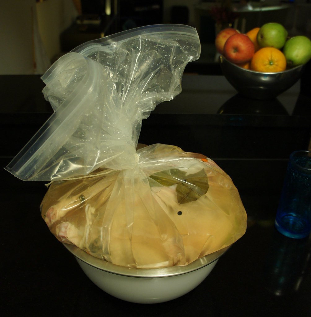 A turkey sealed in a clear plastic brining bag full of golden brine with bay leaves and peppercorns, set in a metal mixing bowl on a dark countertop