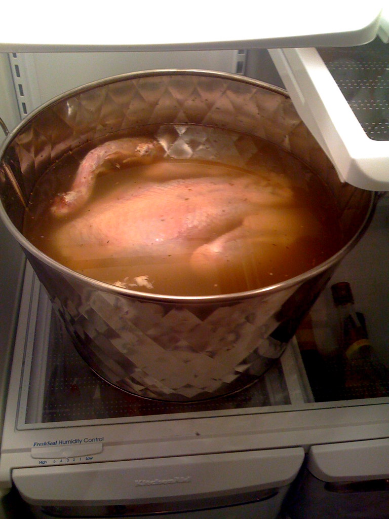 A raw whole turkey fully submerged in amber-colored brine flecked with herbs, inside a large stainless steel tub sitting on a refrigerator shelf